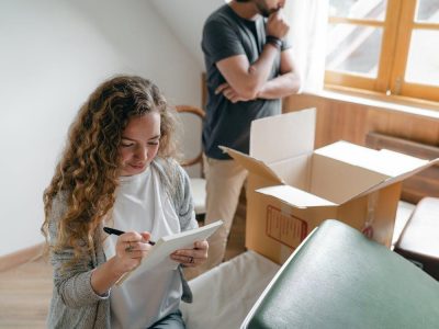 Pensive couple taking notes in bedroom near window while packing stuff in carton box to move into new apartment